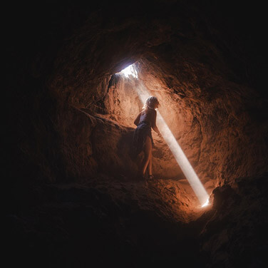 a woman stands in a dark cave with a beam of light coming through the ceiling. her body language looks happy and lively